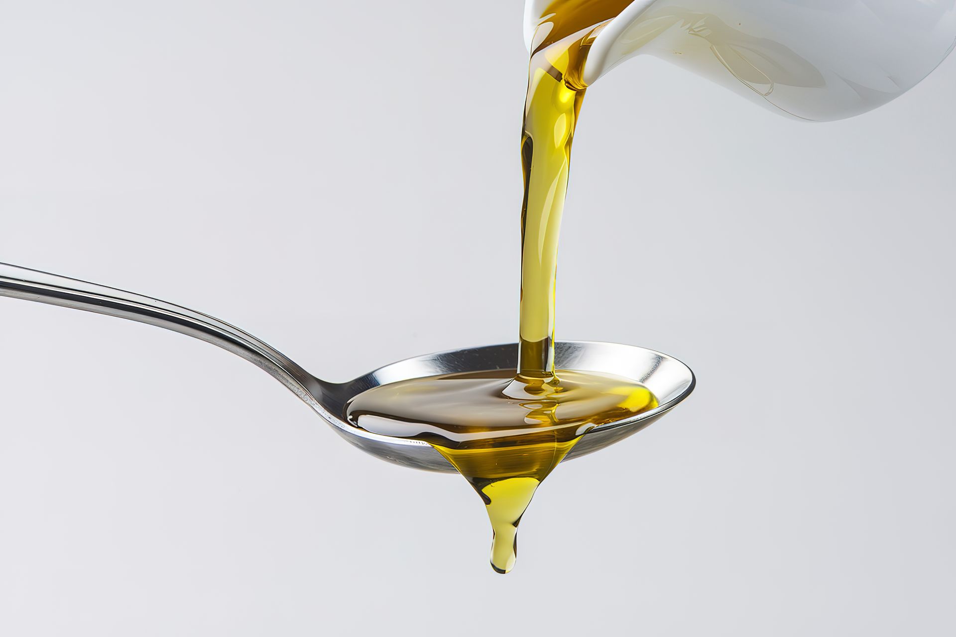 Olio d'oliva versato da un contenitore di vetro in un cucchiaio su sfondo bianco.