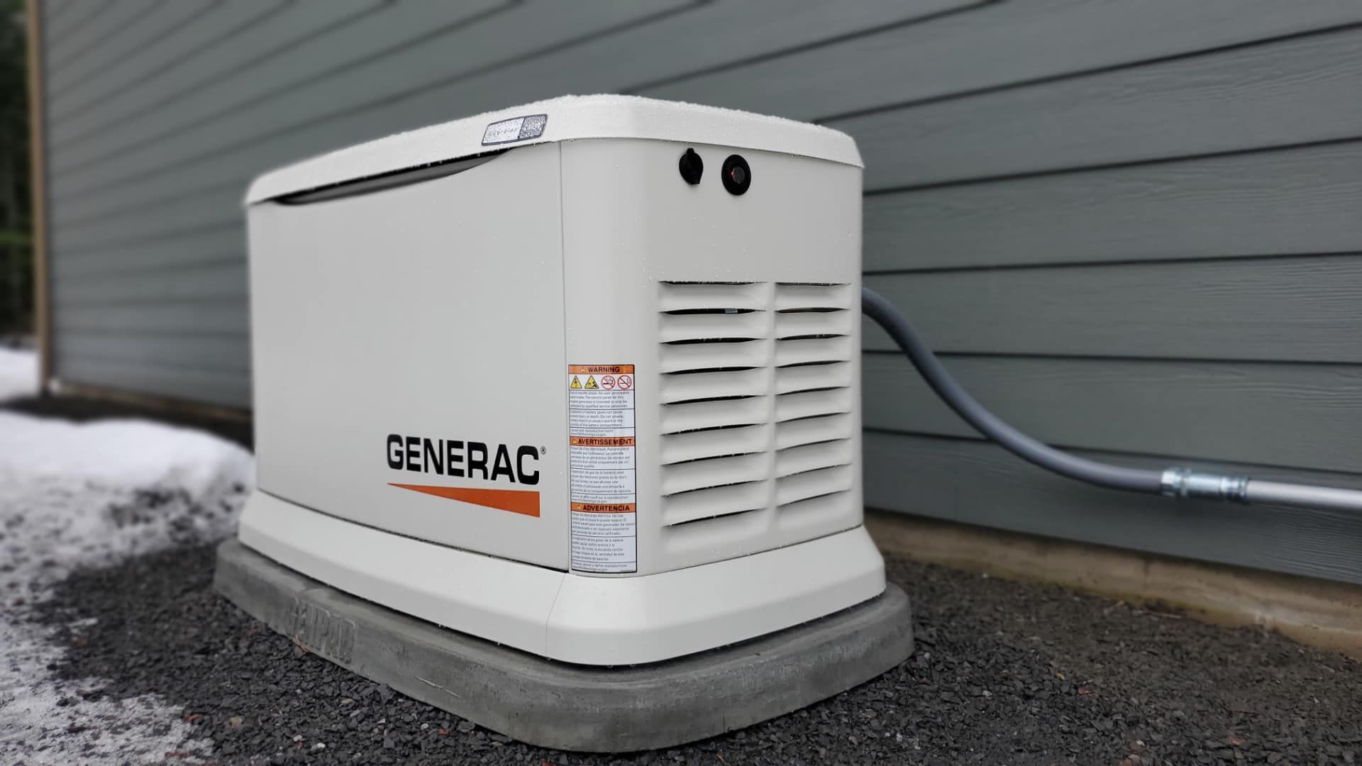 White Generac standby generator beside a house, sitting on a concrete pad near snow-covered ground.
