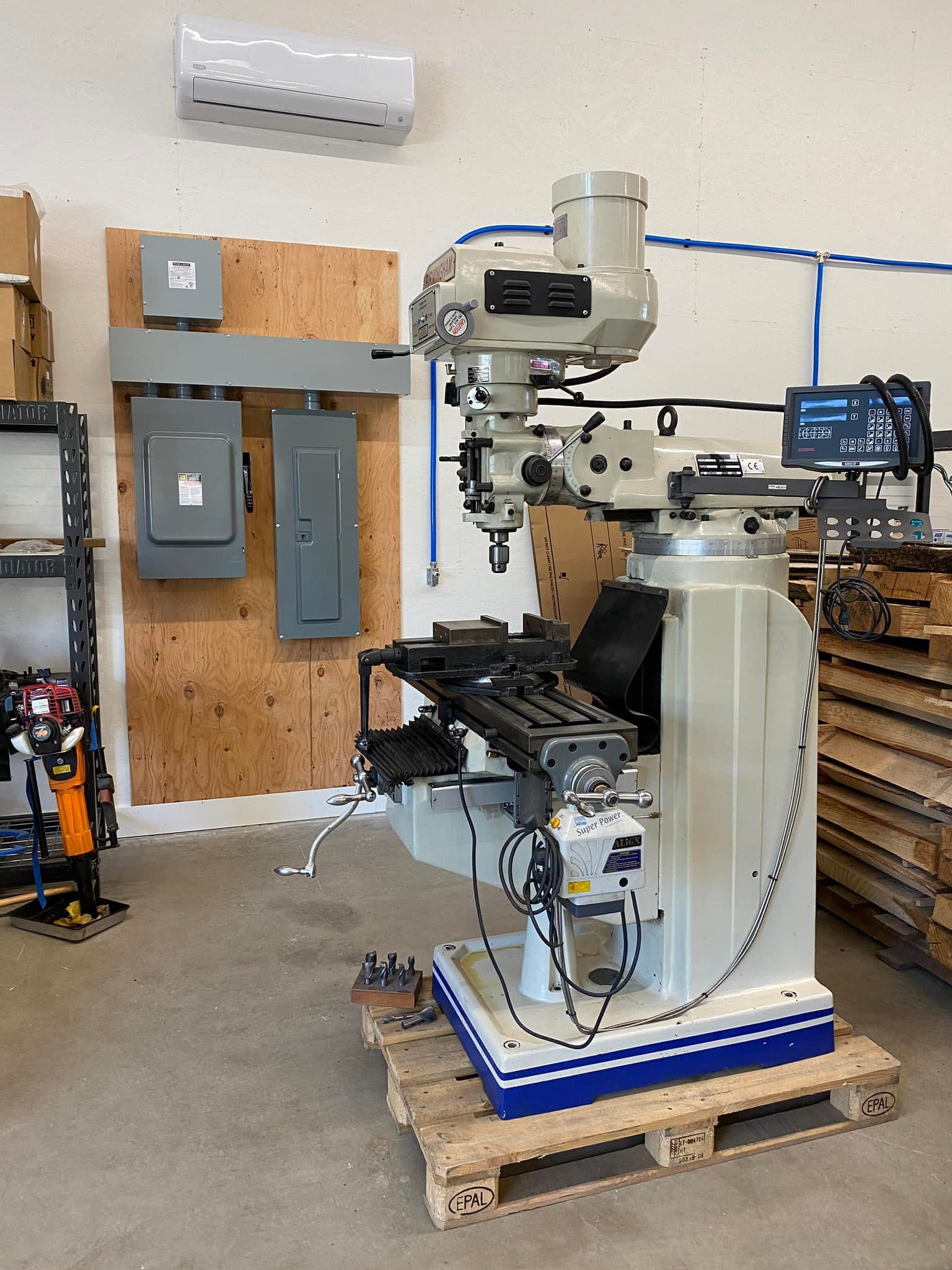 Industrial machine setup on a wooden pallet in a workshop, with wall-mounted electrical boxes and a small monitor.