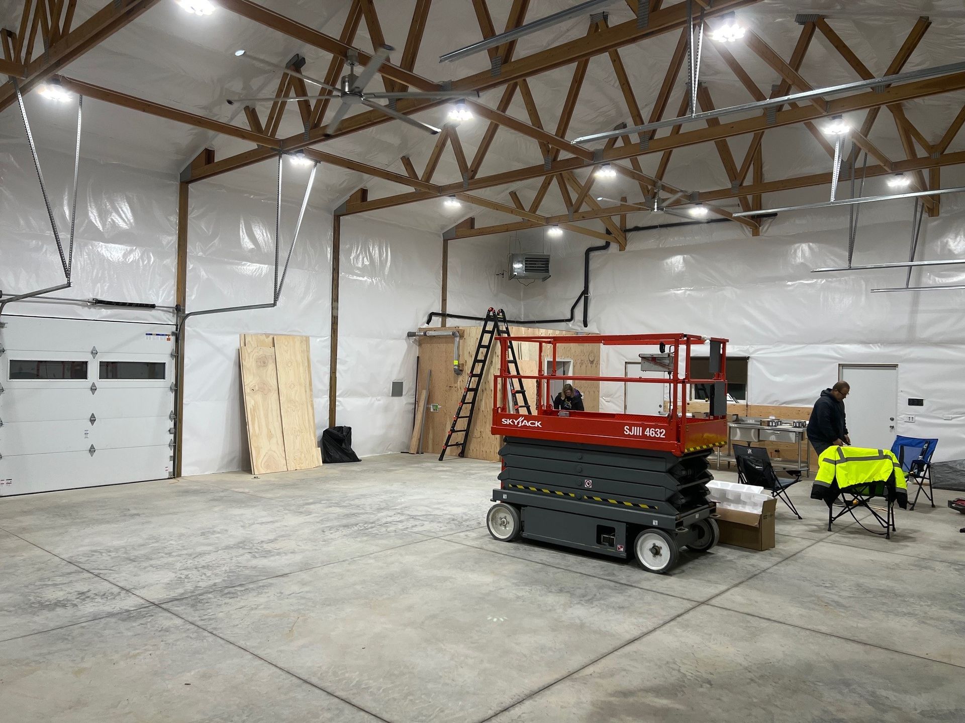Empty industrial warehouse with a red scissor lift, ladder, and two workers near the back wall.