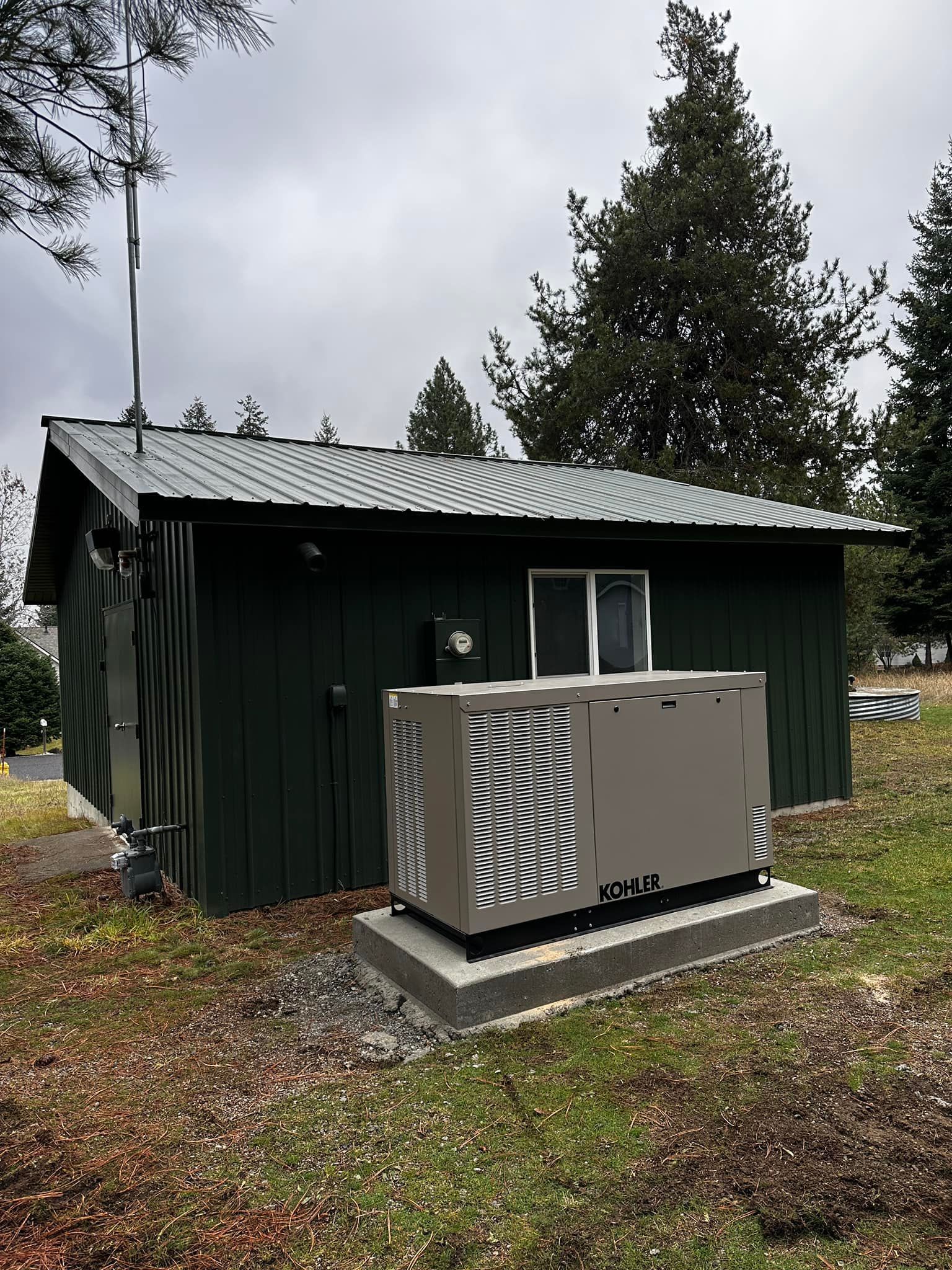 Small dark green utility shed with a metal roof beside a wooded yard, with two large outdoor units in front.