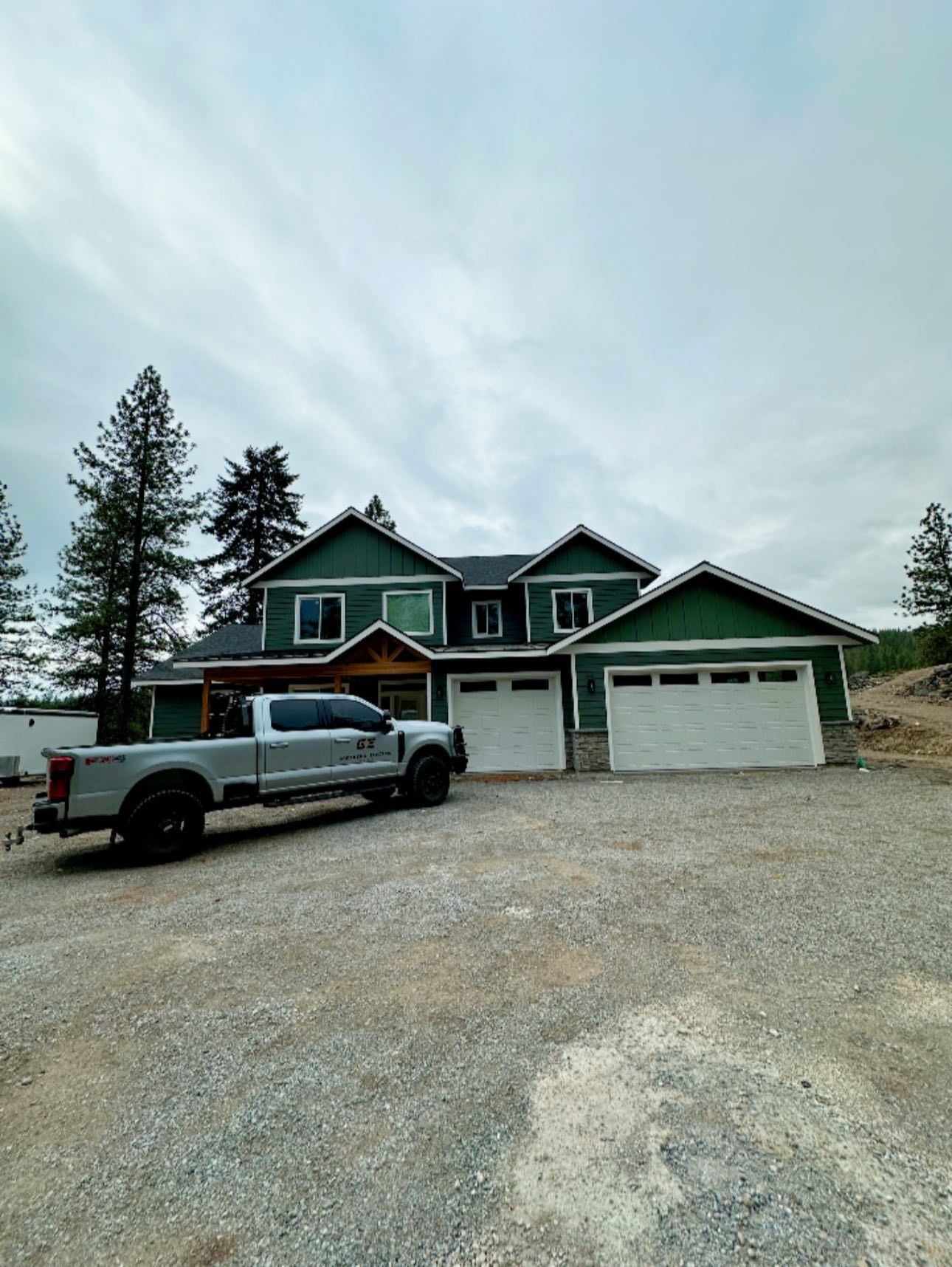 Green house with three garage doors and a white truck parked on a gravel driveway under a cloudy sky
