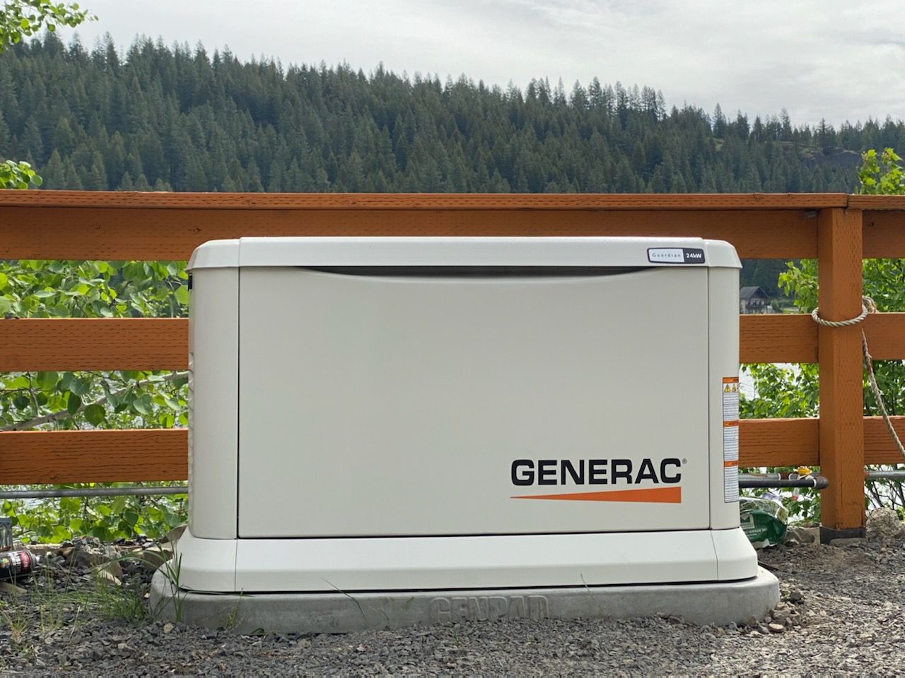 White Generac backup generator on a concrete pad beside a wooden fence, with forested hills in the background