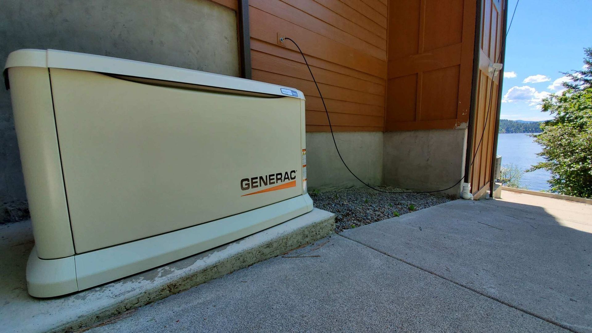 Cream-colored Generac backup generator beside a house on a sunlit concrete patio, near a lake.