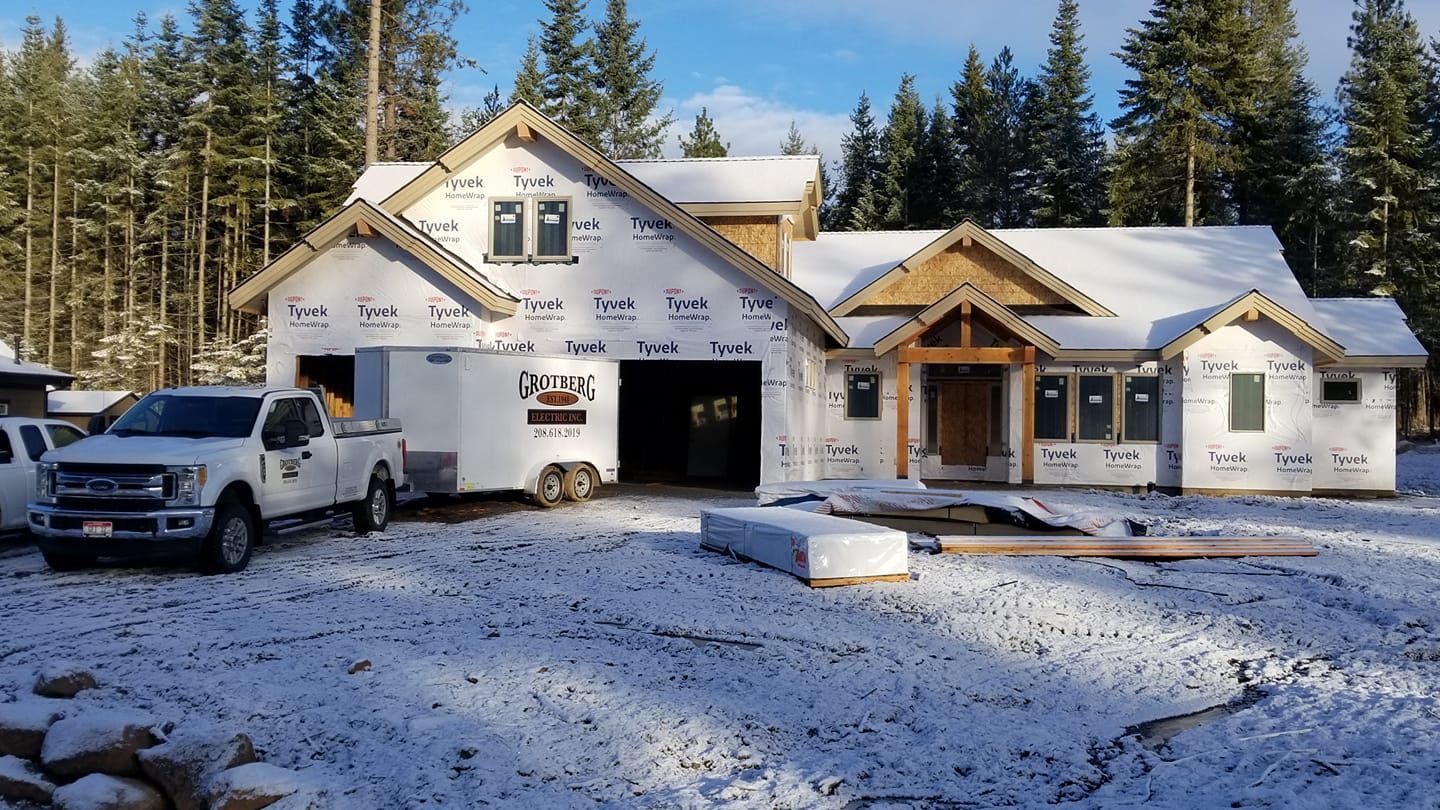 Snow-covered house under construction with a white pickup truck parked out front, pine trees in the background