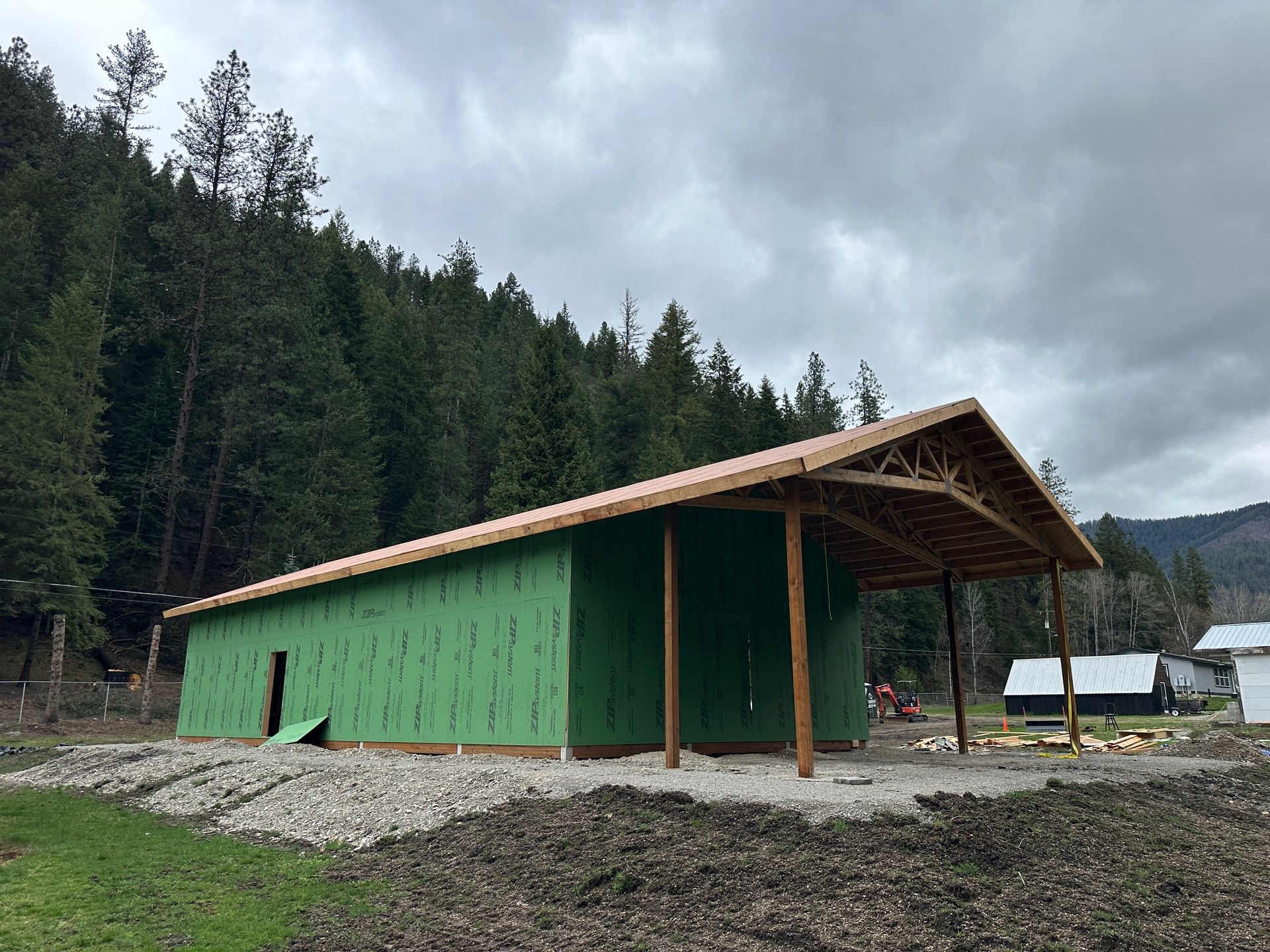 Green cabin with a sloped roof on a gravel site beside a forest, under cloudy skies