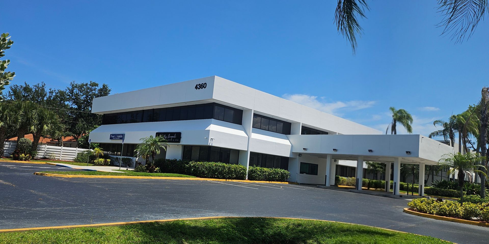 Modern white office building with dark windows, surrounded by palm trees and a sunny blue sky