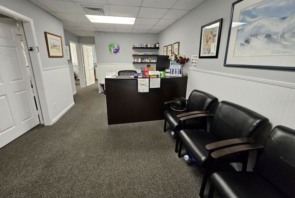 Reception area of a medical office with a dark desk, shelves of medications, and a waiting chair.