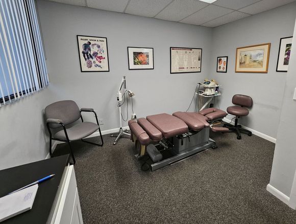 Chiropractor's treatment room with a padded table, equipment, and a stool. Gray walls and medical charts.