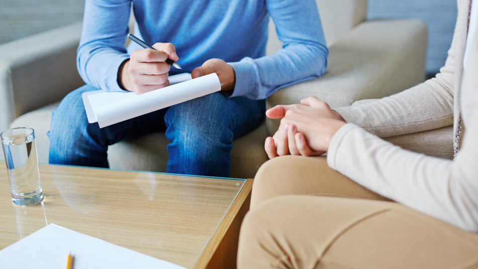 Therapist taking notes, seated across from client with hands clasped, in a light-filled office.