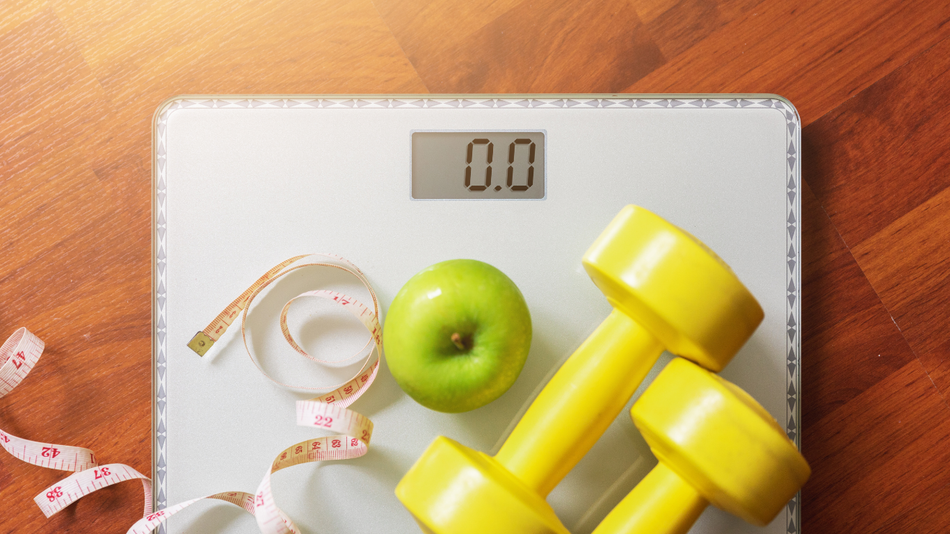 Weight scale with 0.0 reading, apple, measuring tape, and two yellow dumbbells on a wood floor.