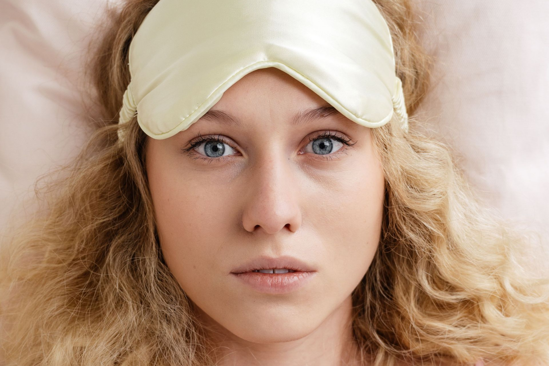 Close-up of a person with blue eyes, curly blonde hair, and a cream sleep mask on their forehead.