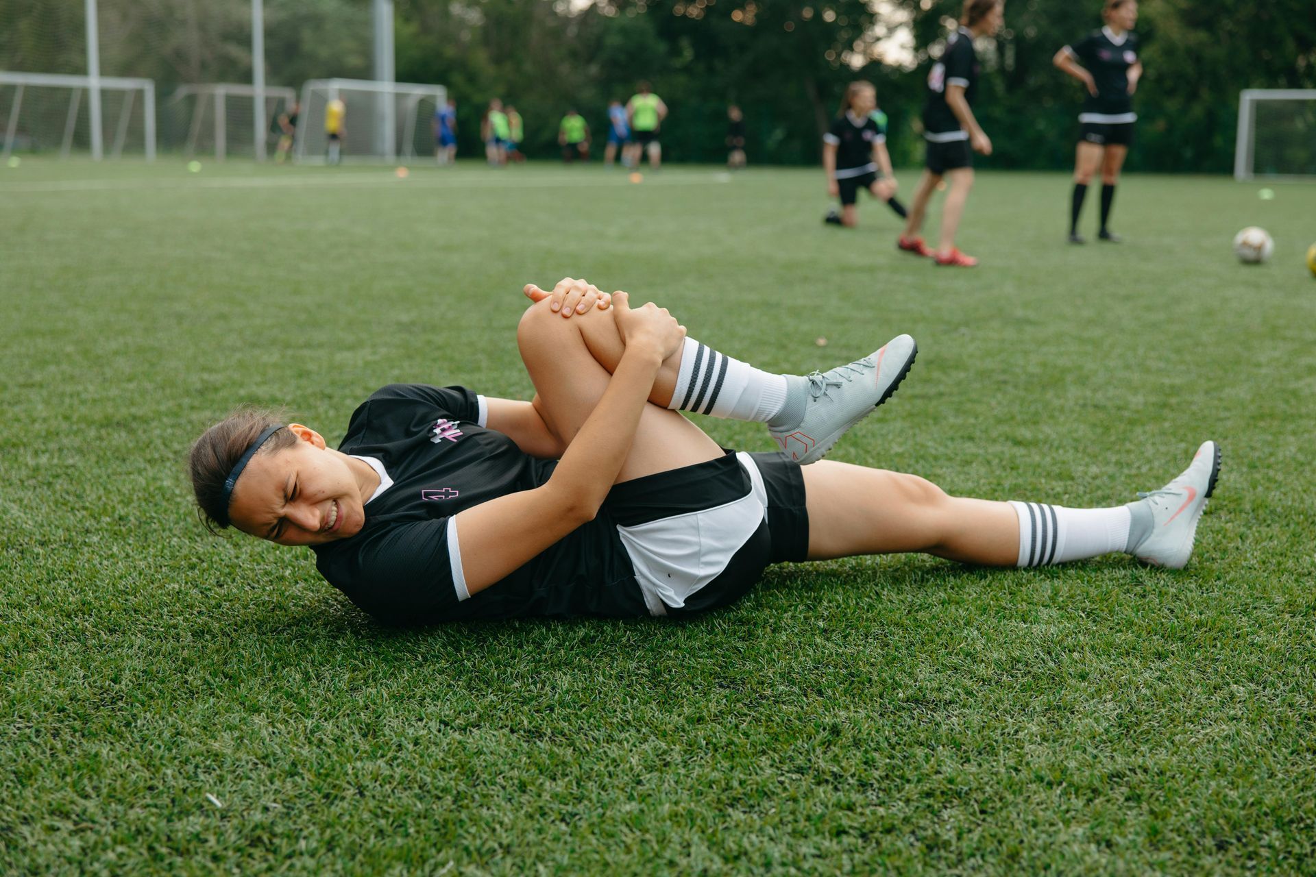 Soccer player lying on grass clutching leg during practice, with teammates in the background