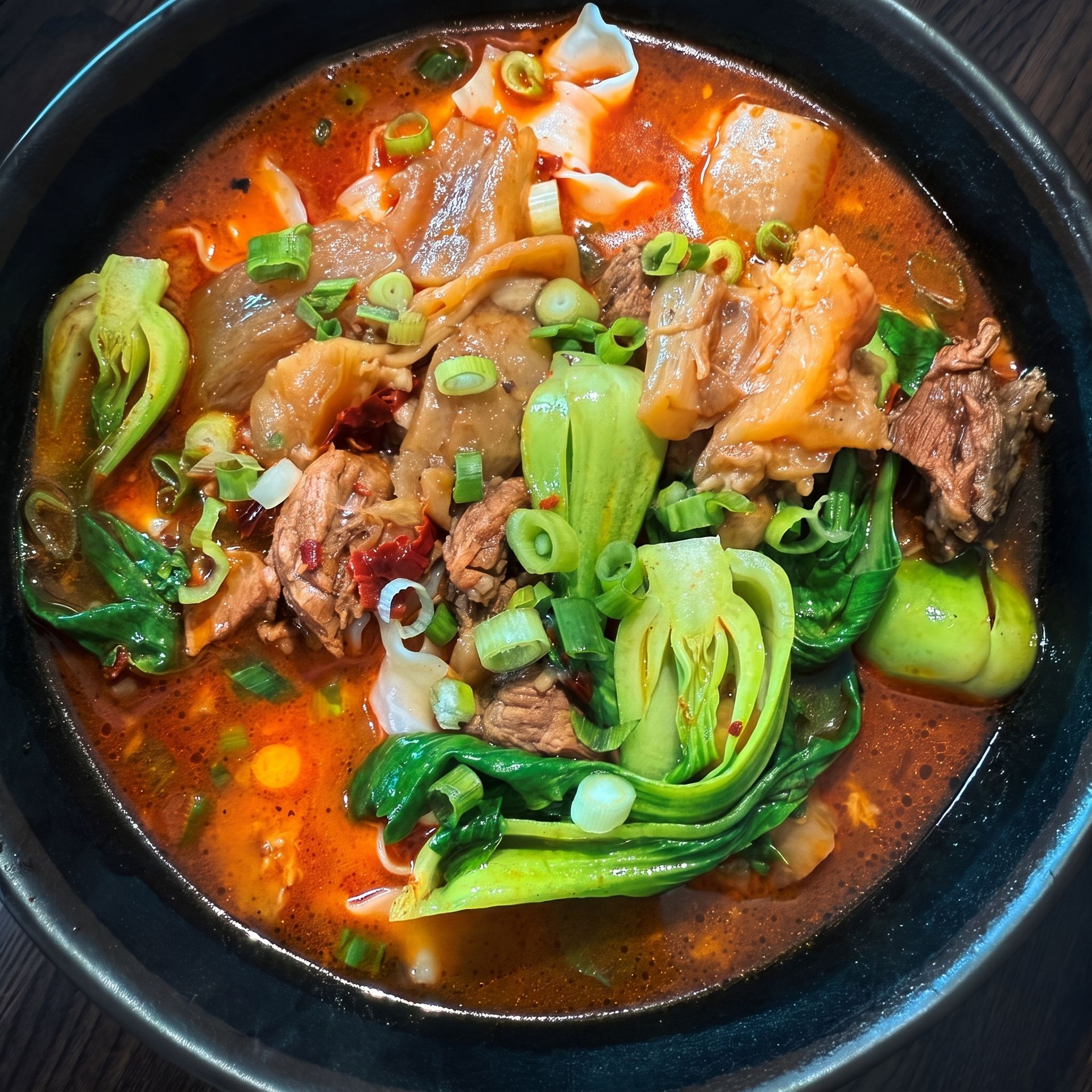 Red spicy soup with meat, tofu skin, and bok choy in a black bowl, topped with green onions