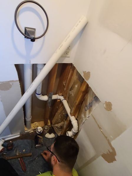 Plumber working under a sink beside exposed pipes and damaged wall in a bathroom area