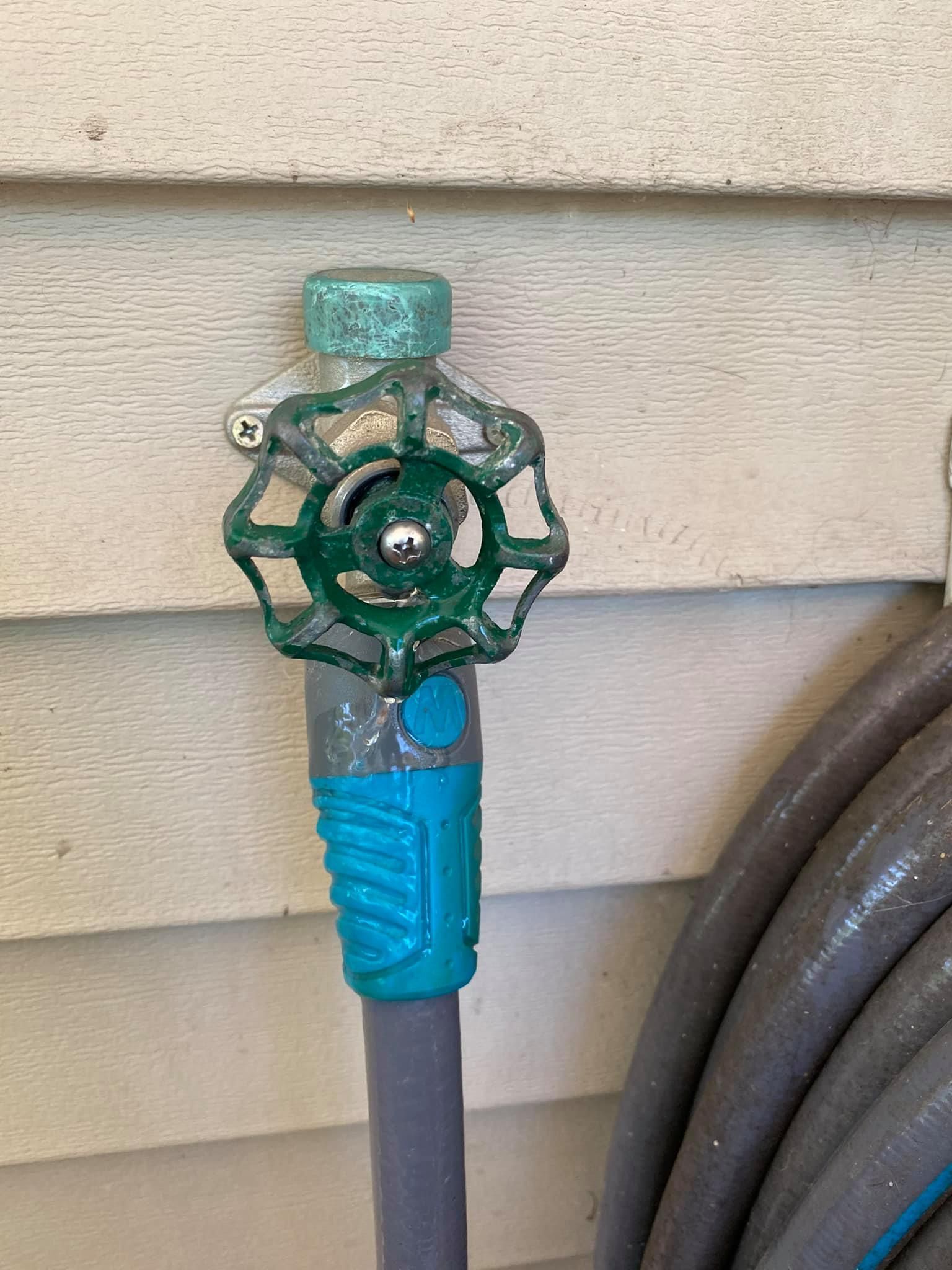 Green garden hose spigot with blue handle attached to a house wall, next to a coiled hose
