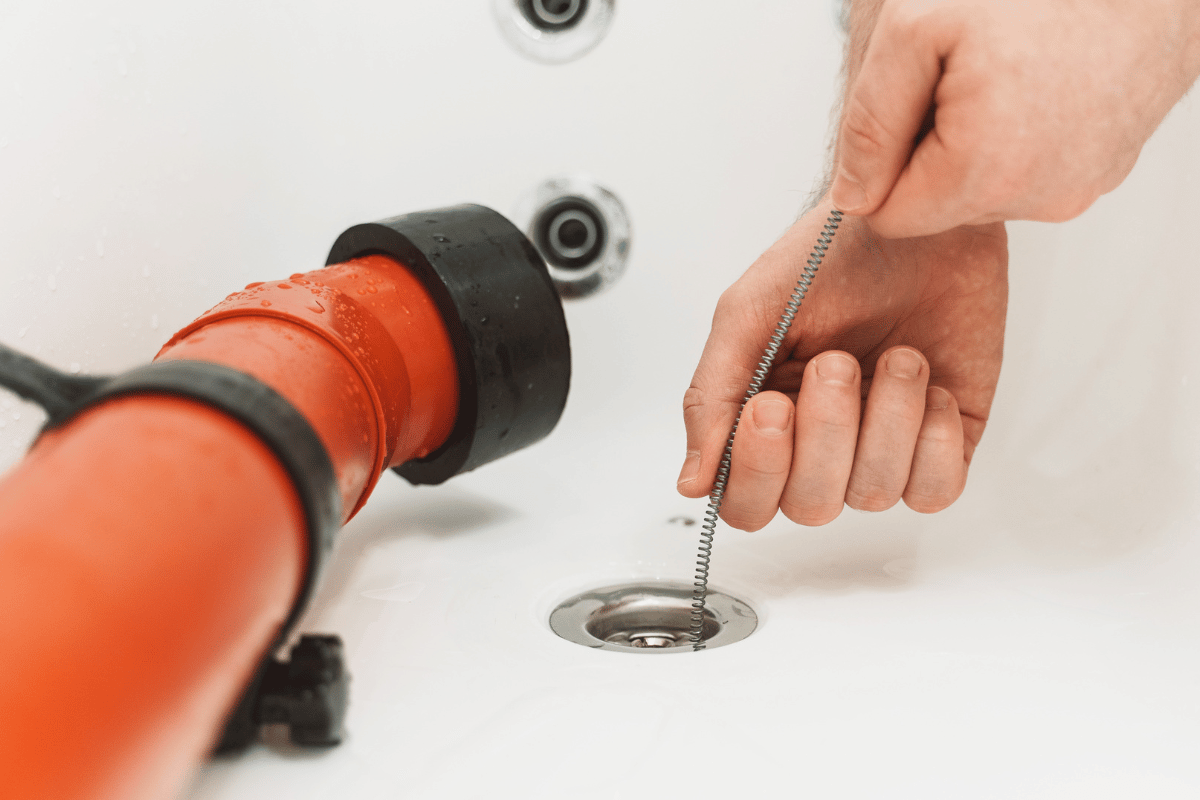 Hand unclogs bathtub drain with a drain snake beside an orange pipe