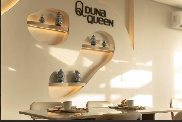 Interior de restaurante com mesas e cadeiras, janela ampla, cores neutras e elementos decorativos na parede.