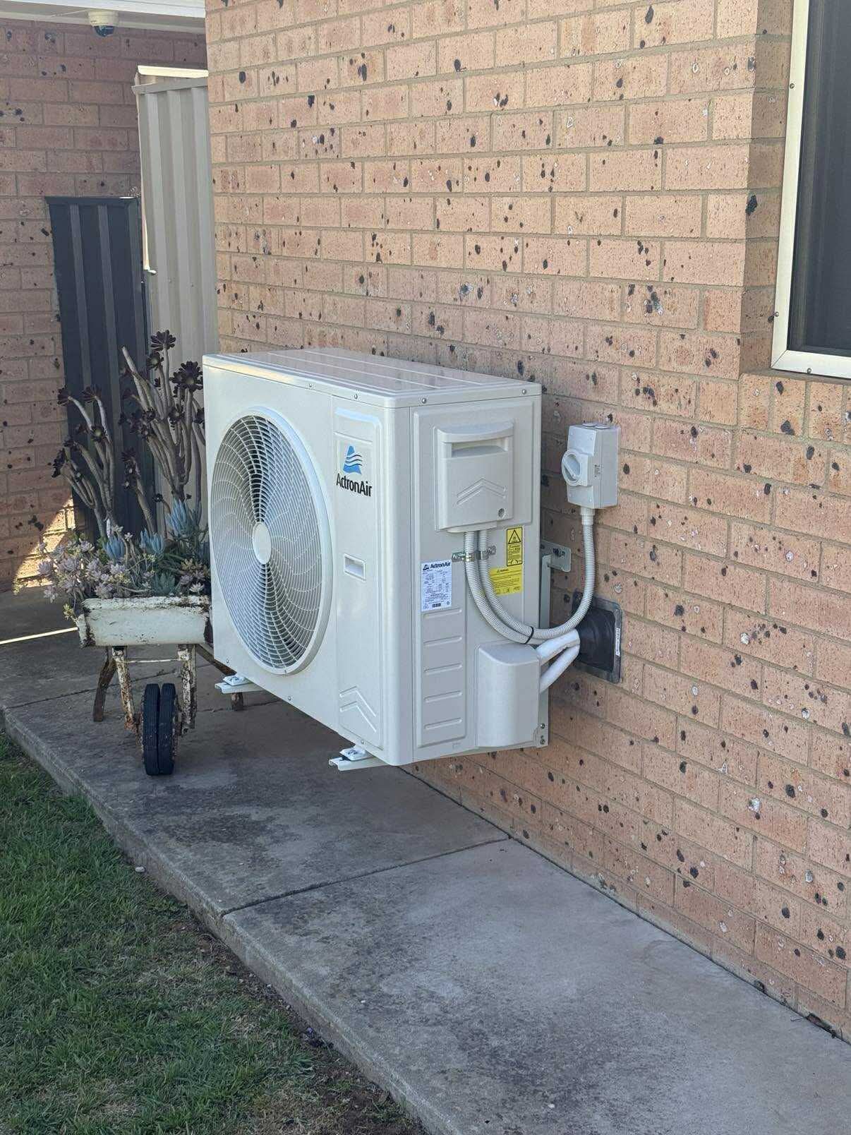 Outdoor air-conditioning unit mounted on a brick house wall beside a window and patio path — Craig Stewart Electrical in East Wagga Wagga, NSW