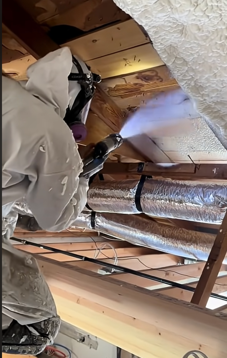 Technician applying spray foam insulation near attic ductwork