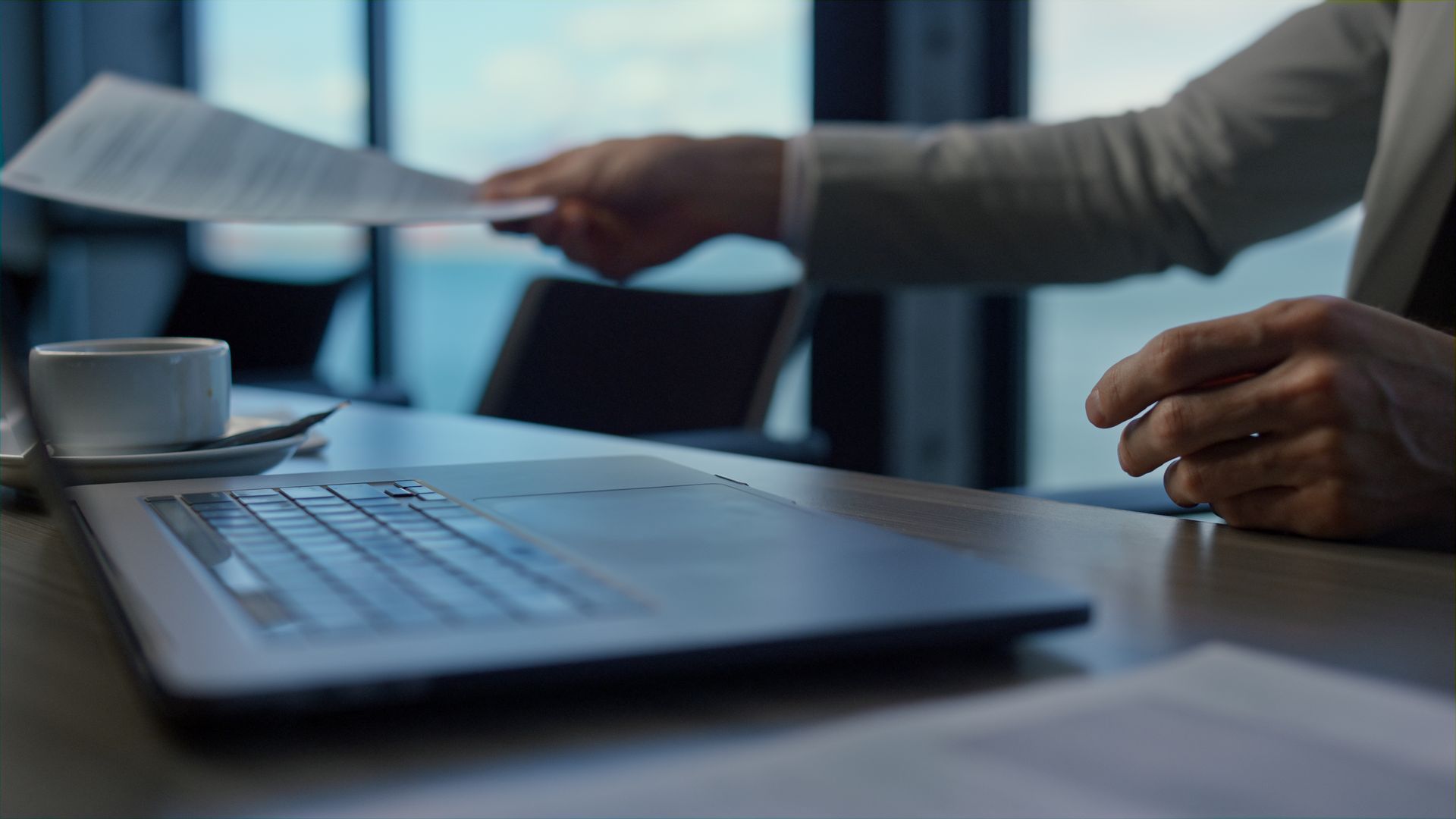 Person handing a document across a desk next to an open laptop and coffee cup in an office setting