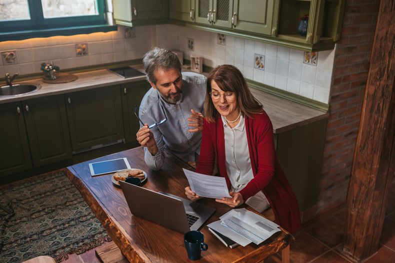 Two people reviewing papers at a kitchen table with a laptop and tablet