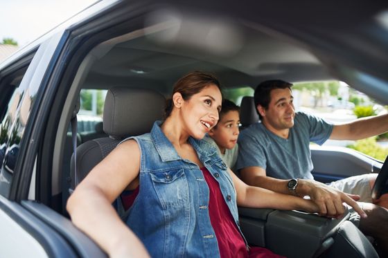 Three people smiling in a car, with a woman in a denim vest in the front passenger seat