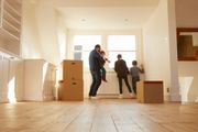 Three people moving boxes in a bright, empty room with wood floors and large windows.
