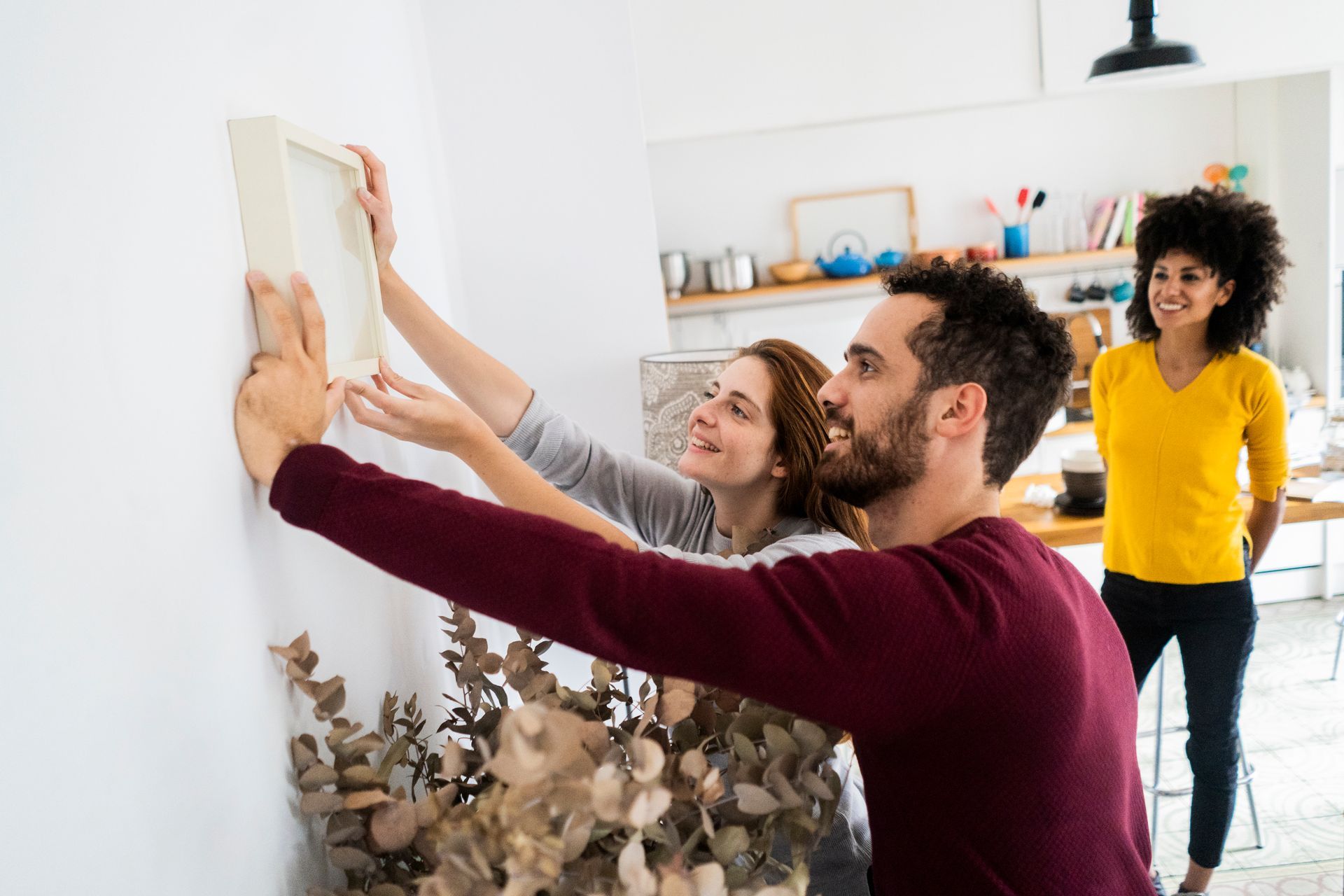 Three people hanging a framed picture on a white wall in a bright room
