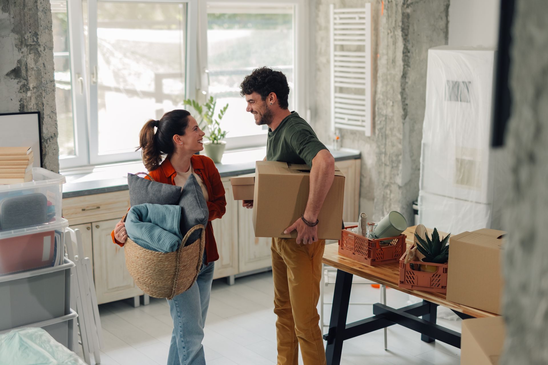 Two people carrying moving boxes in a bright room near a window and table.