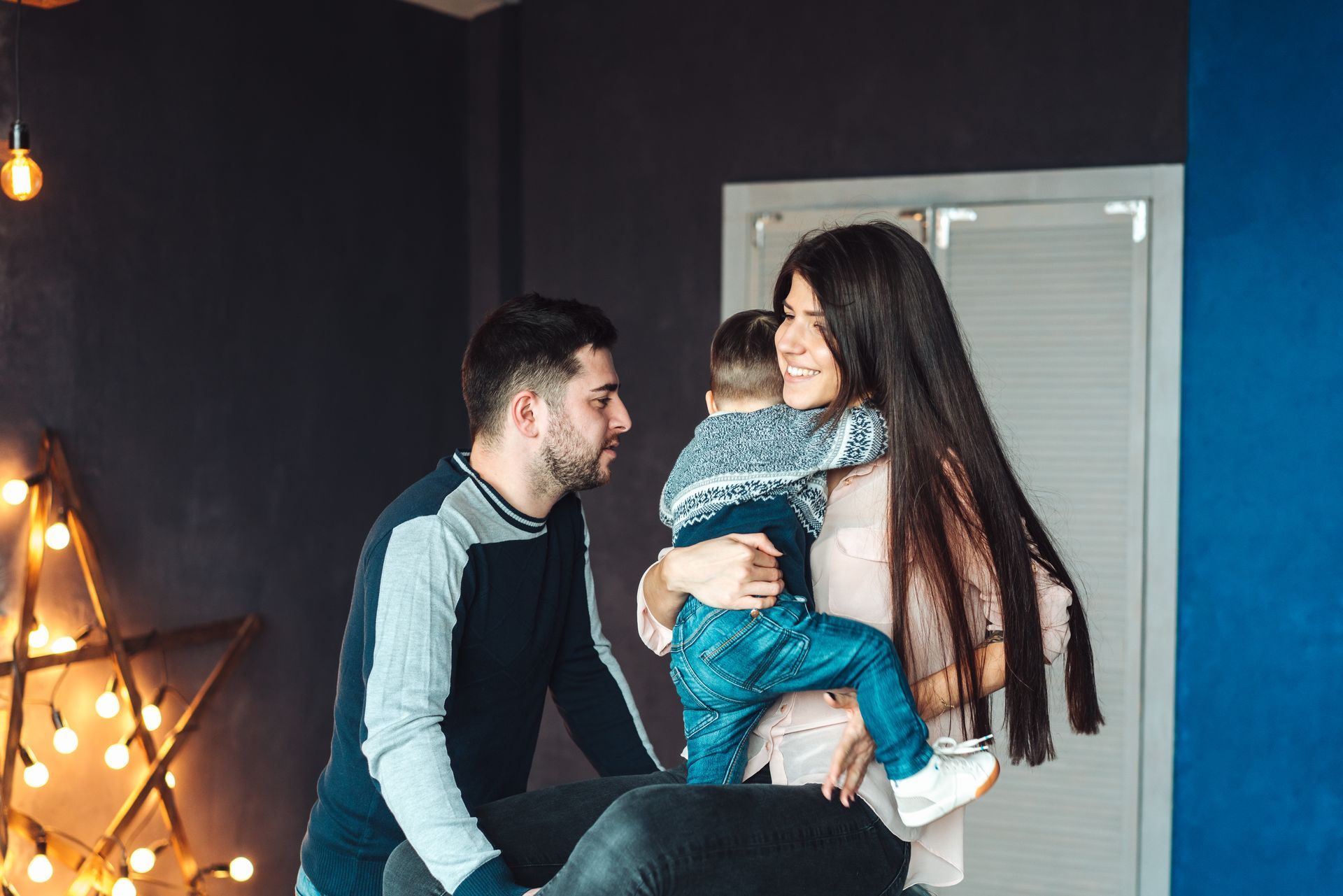 Man and woman smiling while holding a child in a cozy room with warm string lights.