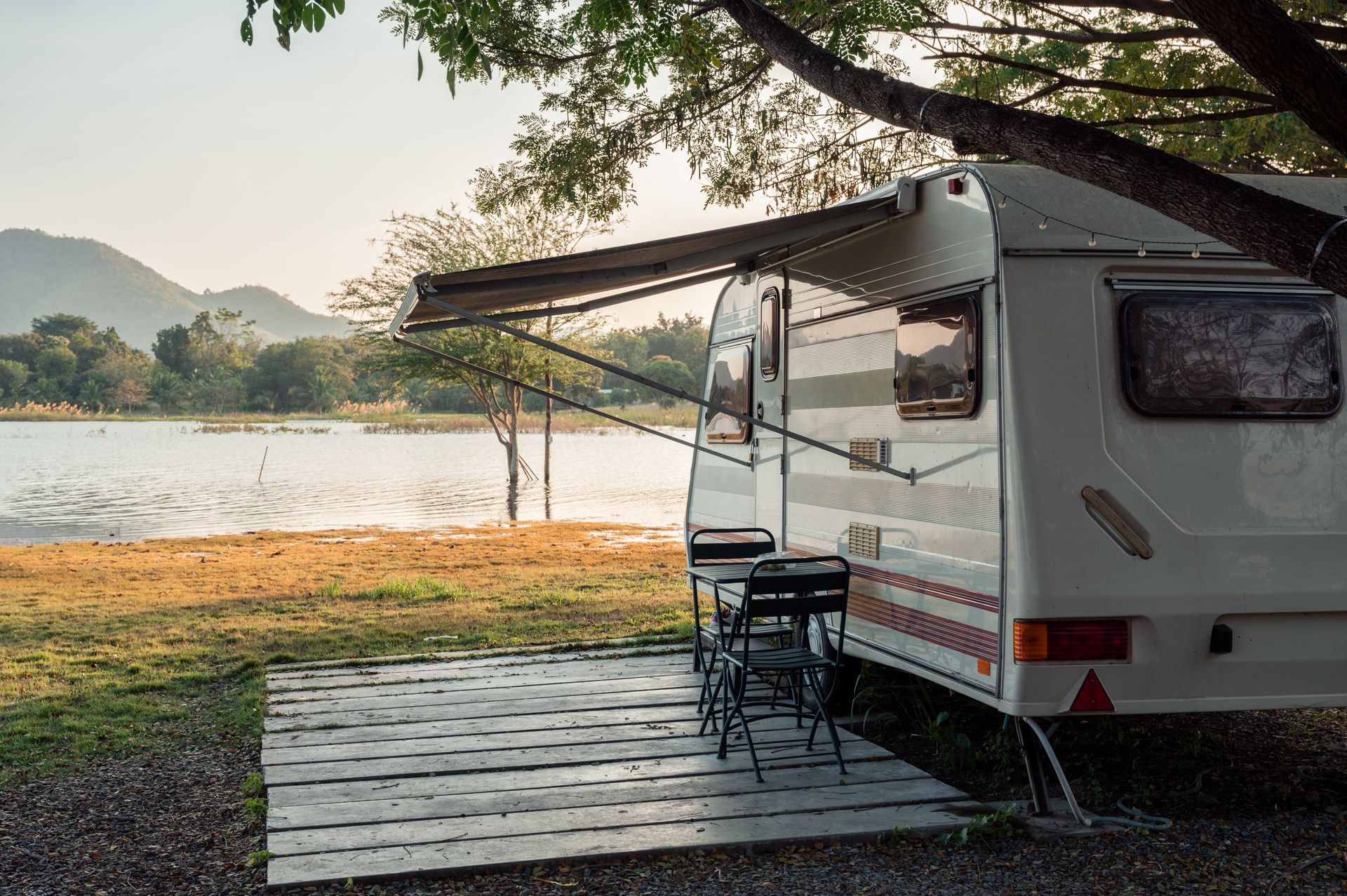 Camper van parked by a lakeside under a tree at sunset, with chairs and a small table outside