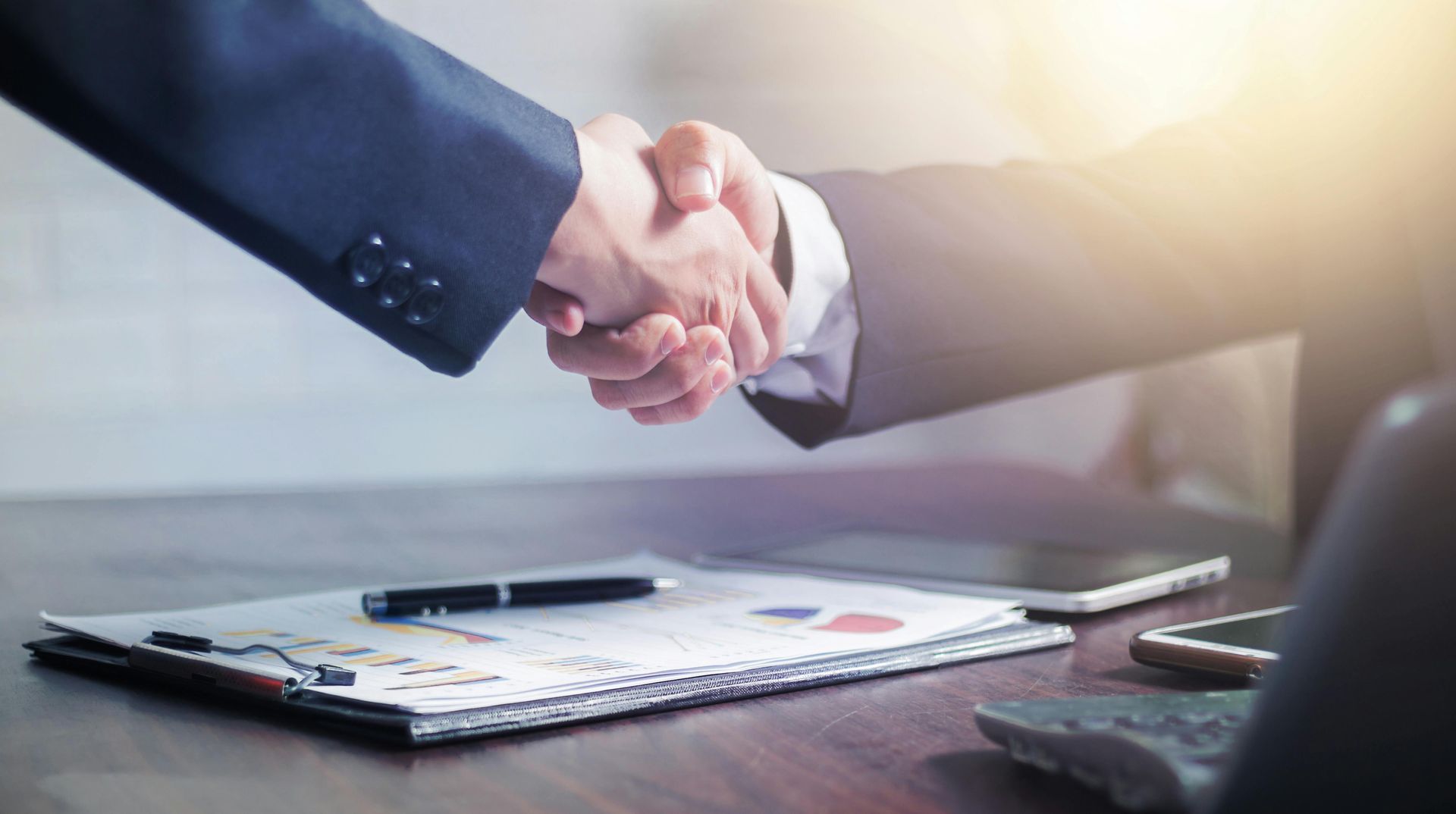 Business handshake over a desk with paperwork, pen, and tablet in a bright office setting