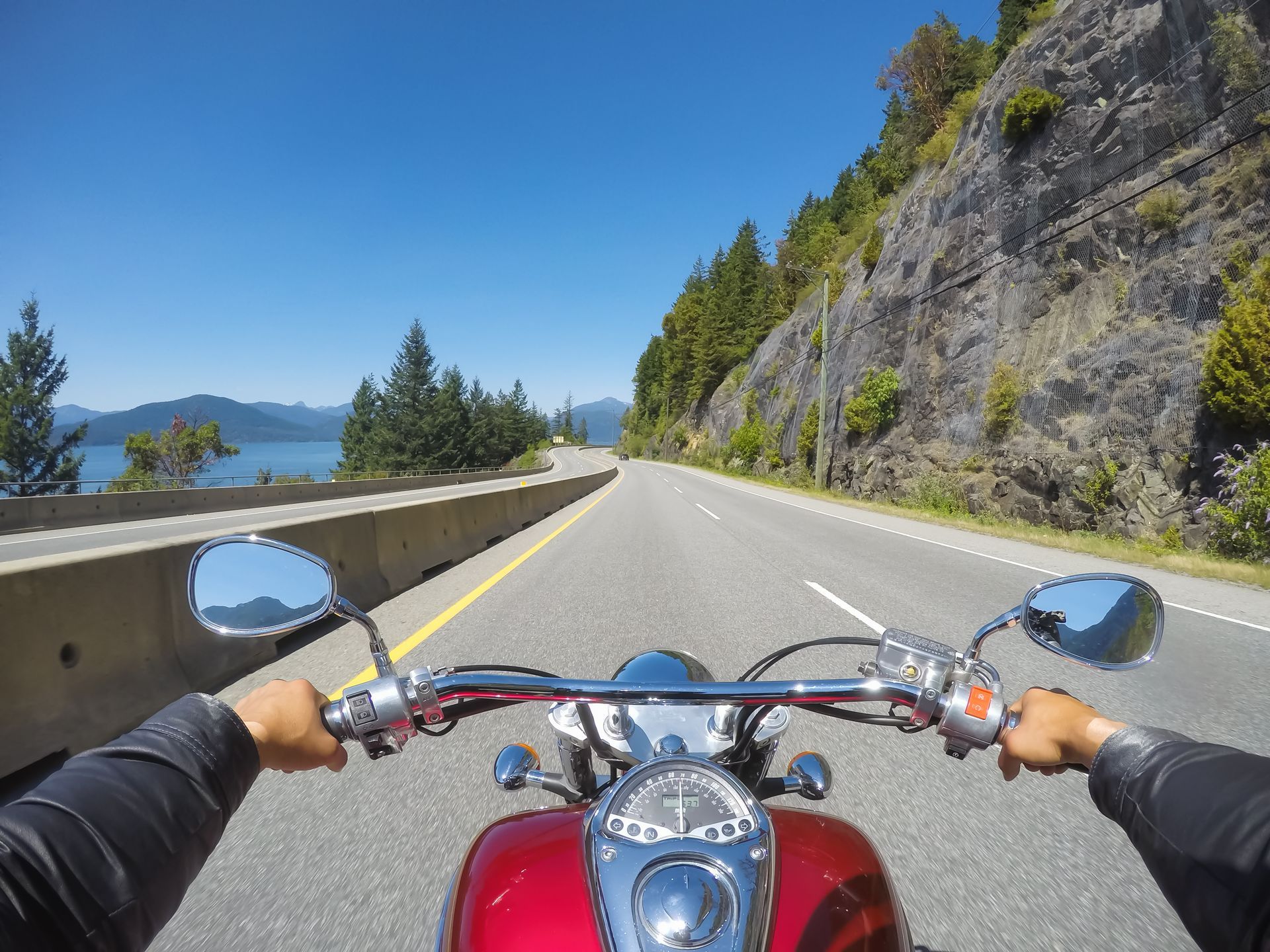 Motorcycle rider on a scenic mountain road beside a lake and rocky cliff under a clear blue sky