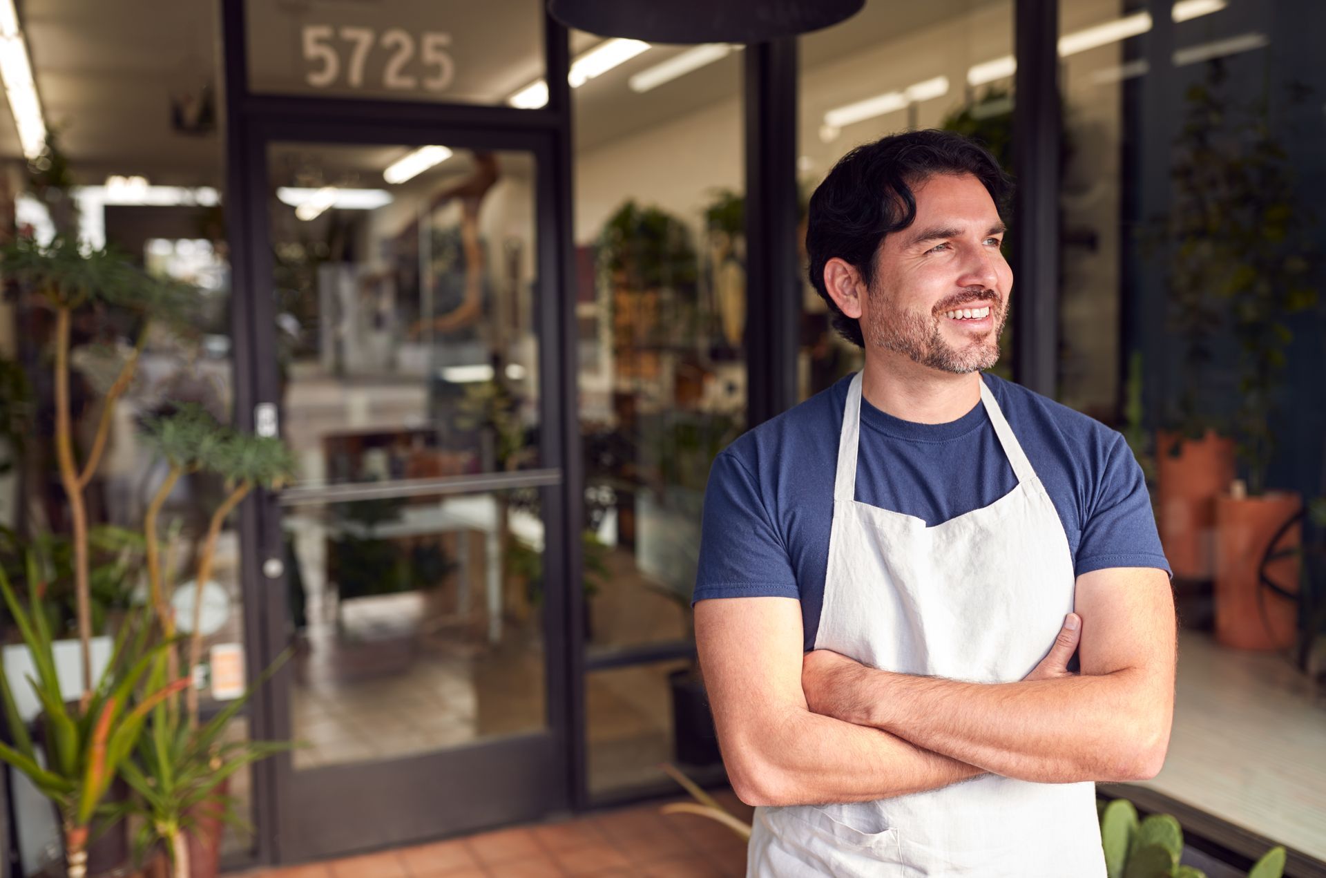 Man standing with arms crossed outside a storefront, wearing a blue shirt and white apron, smiling.