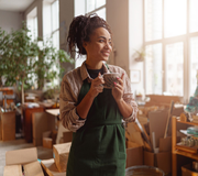 Smiling person in a green apron holding a mug in a sunlit workshop with boxes and plants.