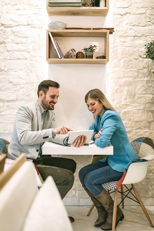 Two people sit at a café table, smiling while looking at a laptop and taking notes.