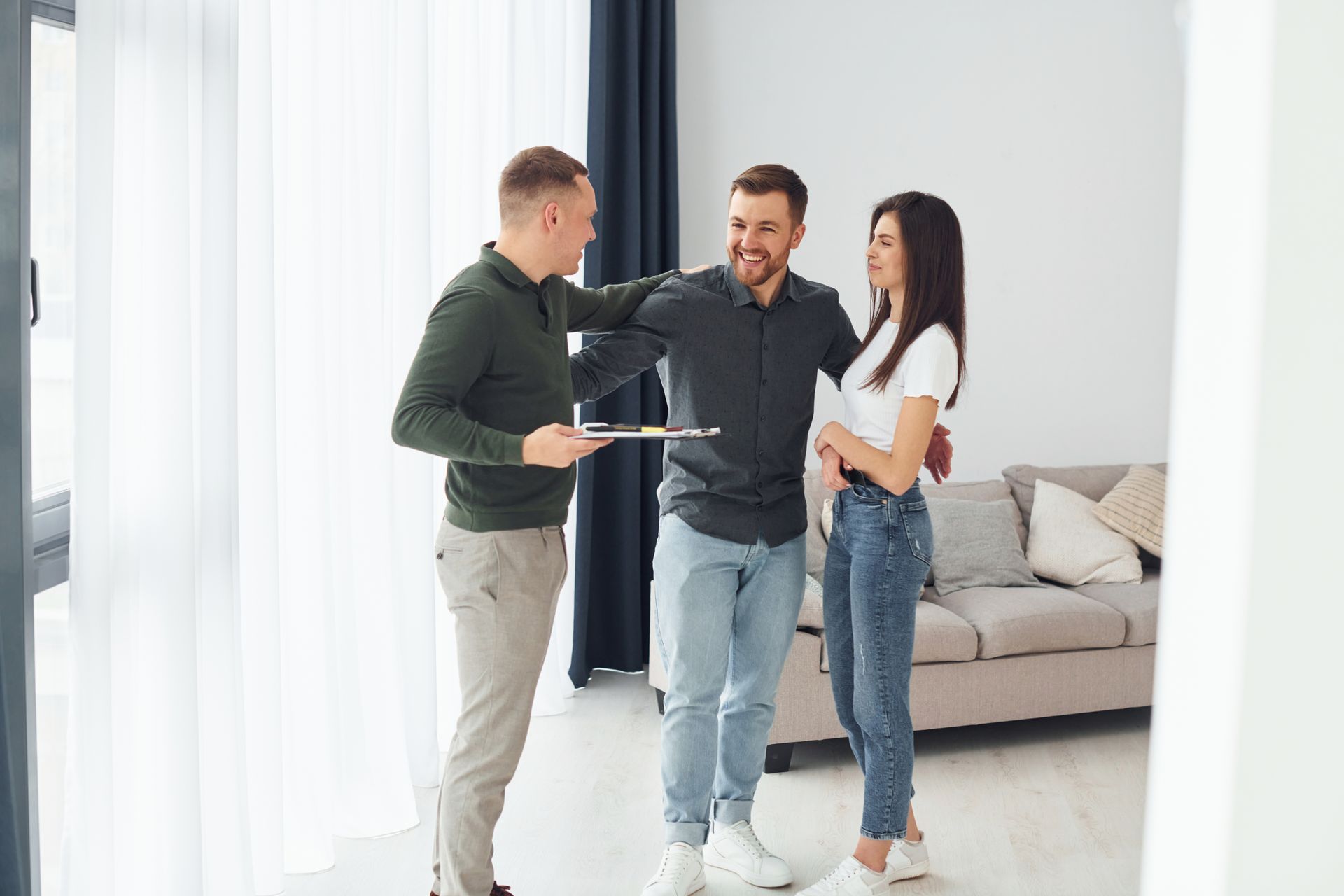 Three people talking in a bright living room, with a sofa and curtains in the background.