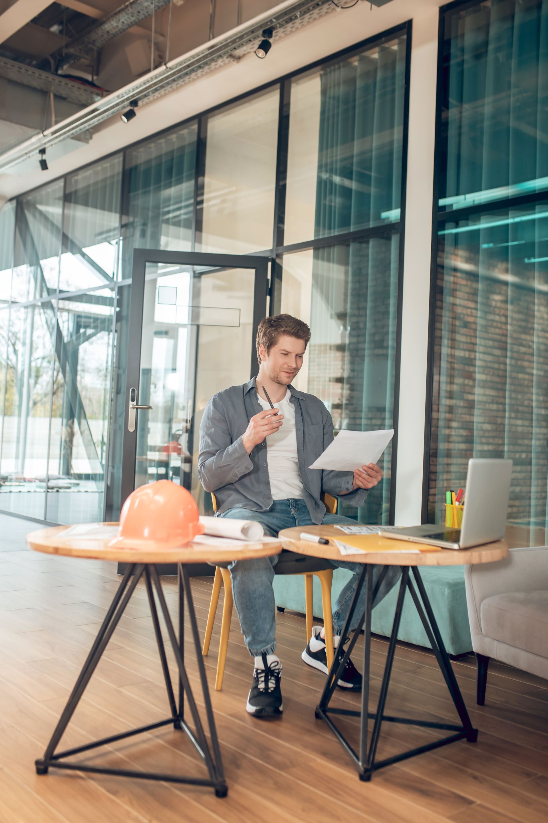 Person reviewing papers at a table in a modern office, with a laptop and orange hard hat nearby.