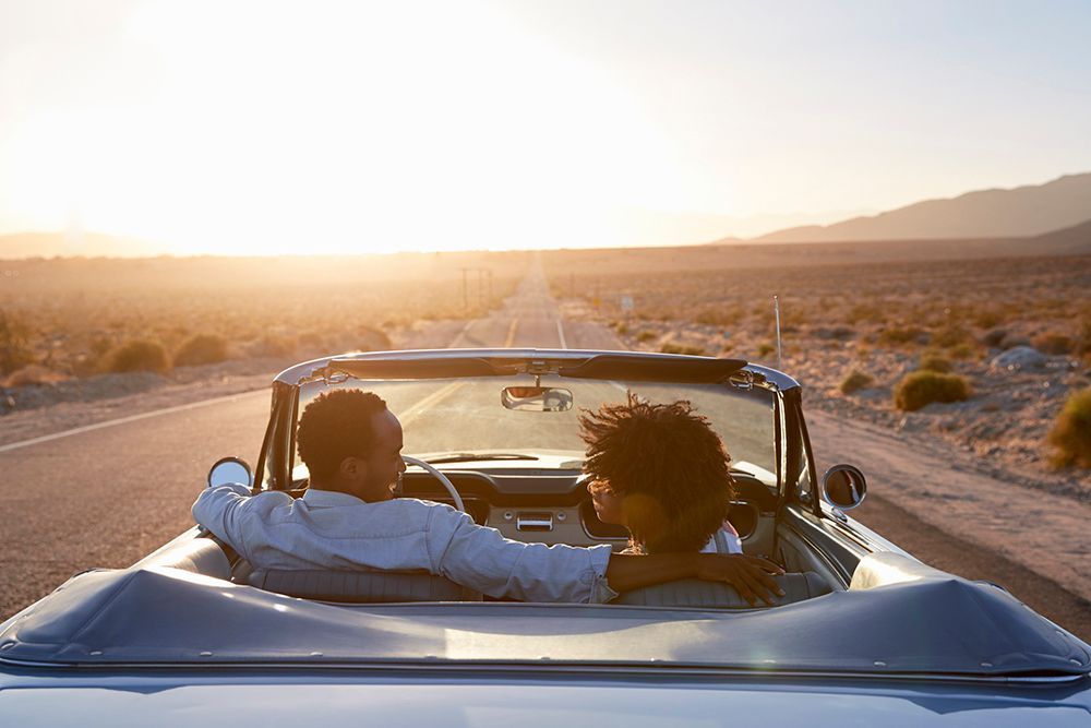 Two people riding in a blue convertible on a desert road at sunset