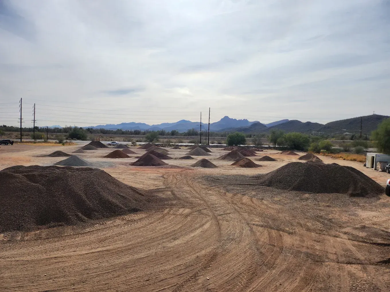 Gravel piles and dirt mounds in an open lot under a cloudy sky, with mountains in the distance