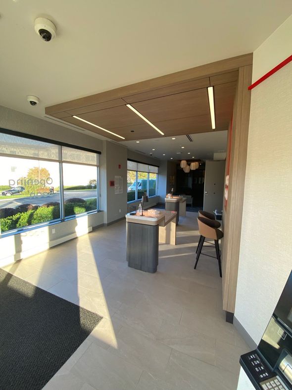 A modern, brightly lit bank interior with tall desks, a window view of trees, and a dark stool on a light tiled floor.