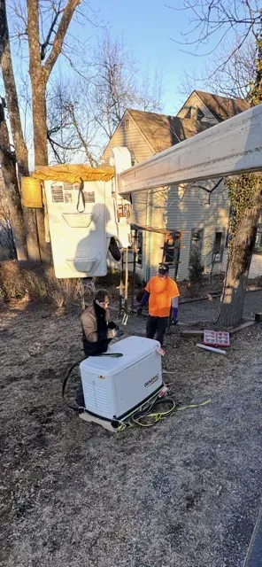 Two workers install a standby generator in a residential yard near a boom lift and a multi-story stone house.