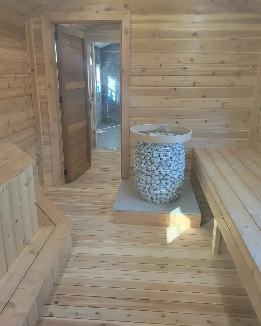 A wooden sauna interior with tiered benches, a central stone heater, and an open doorway leading to an exterior space.