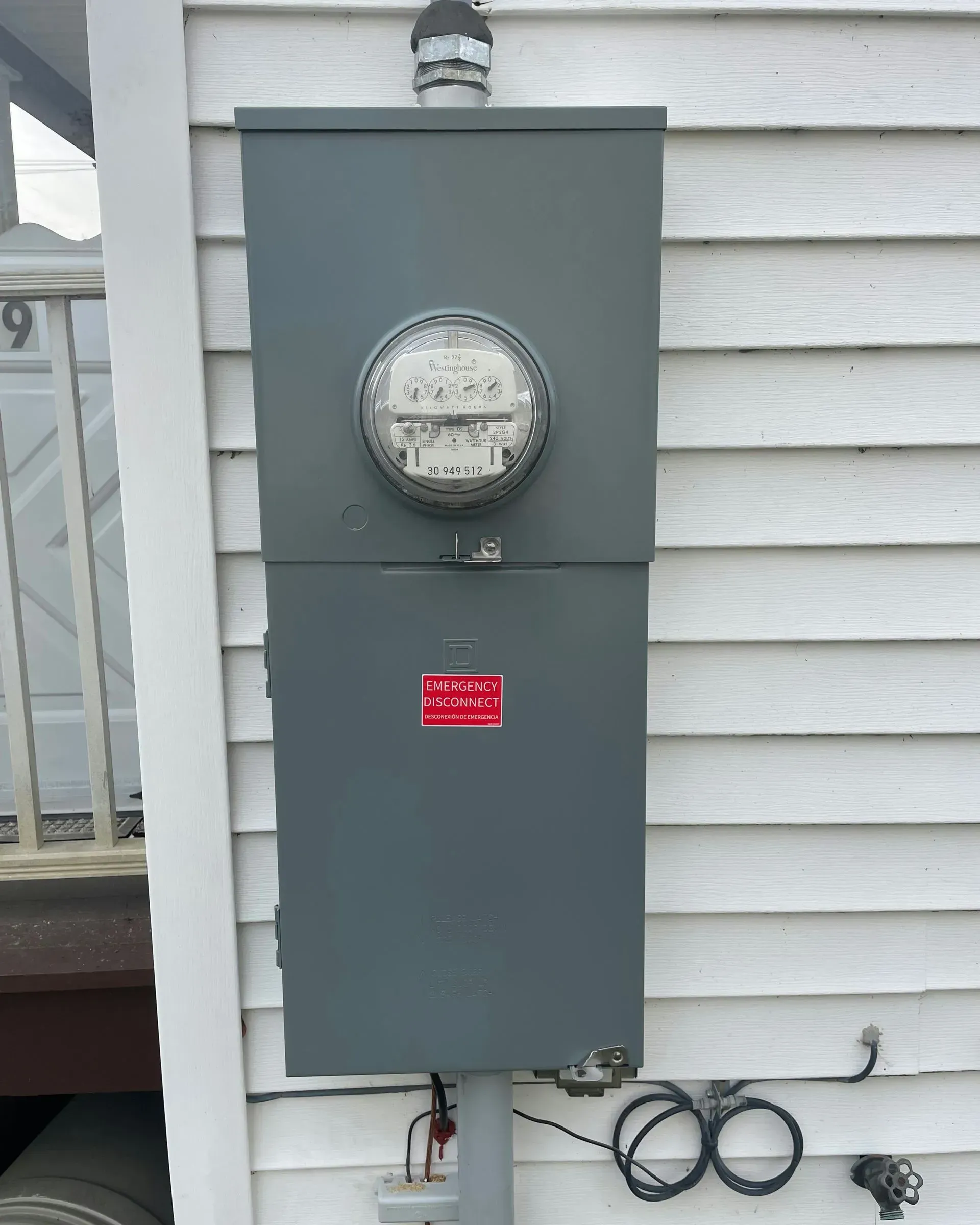 A grey electrical meter box mounted on white vinyl siding, featuring a circular analog meter and a red warning label.