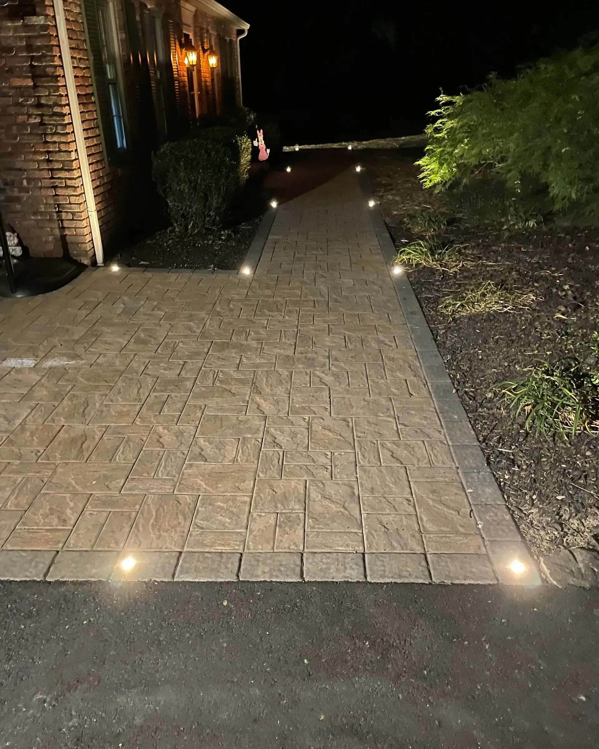 A paved walkway at night, illuminated by soft, recessed ground-level lights along its edges leading toward a house.