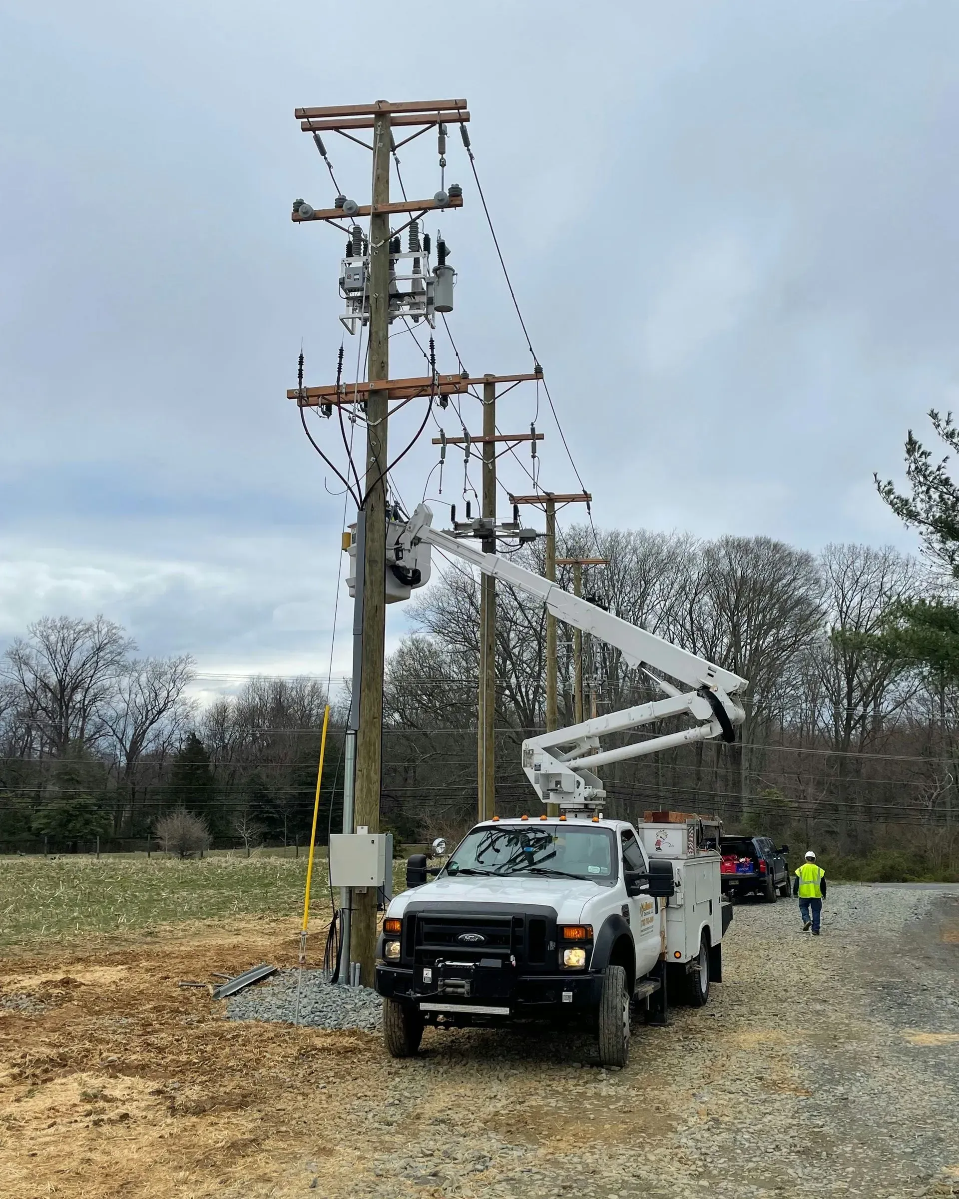 A utility truck with an extended bucket lift parked near power poles in a field, with a worker walking on a gravel path.