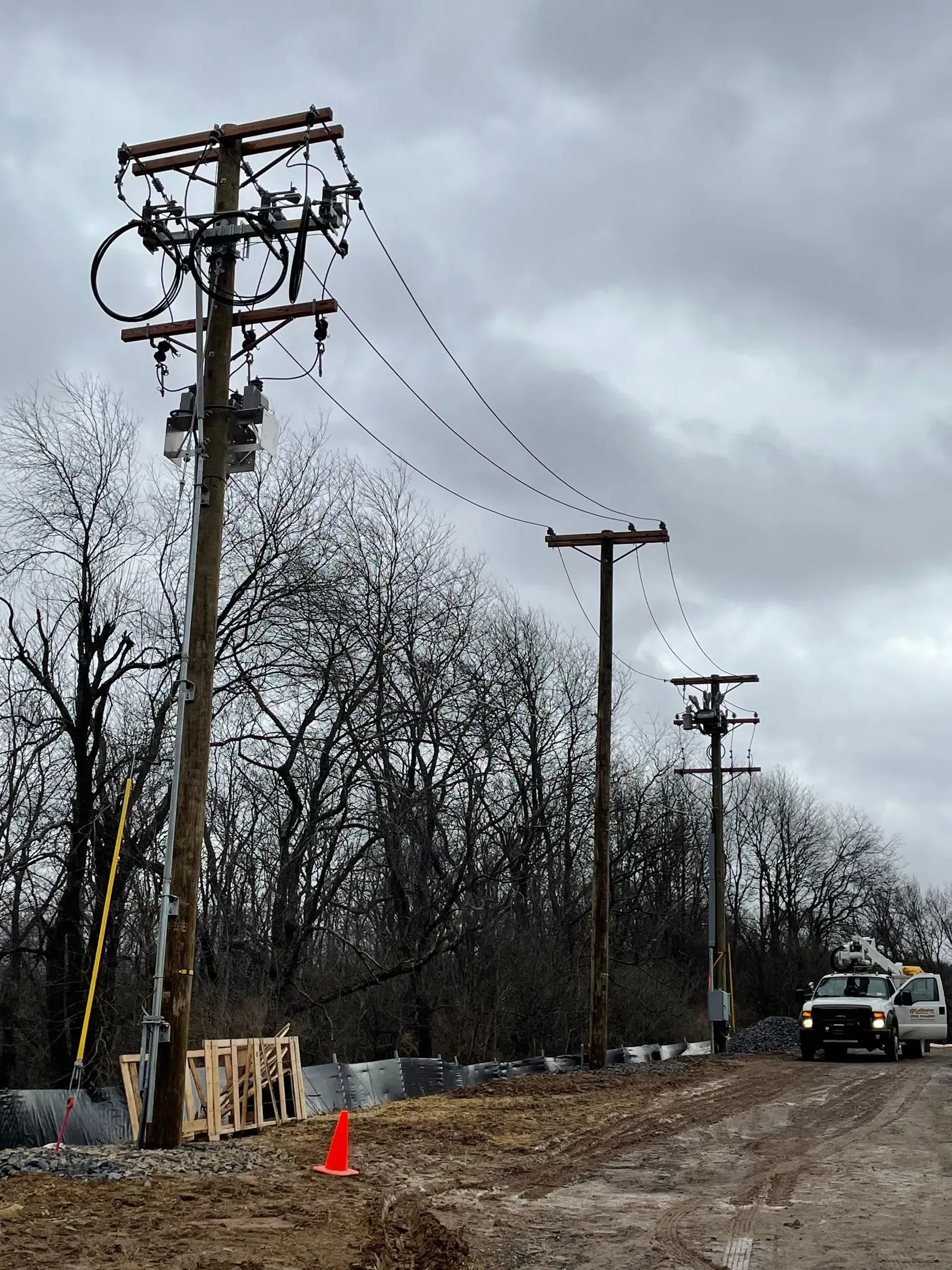 Utility poles with power lines stretching along a road, with a service truck parked nearby on a cloudy day.