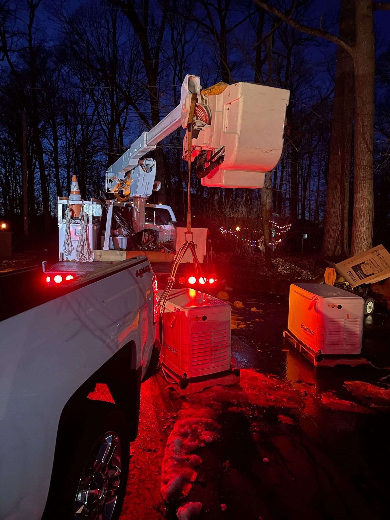 A utility truck with an extended bucket lift operates in a wooded area at twilight, with equipment glowing under red lights.