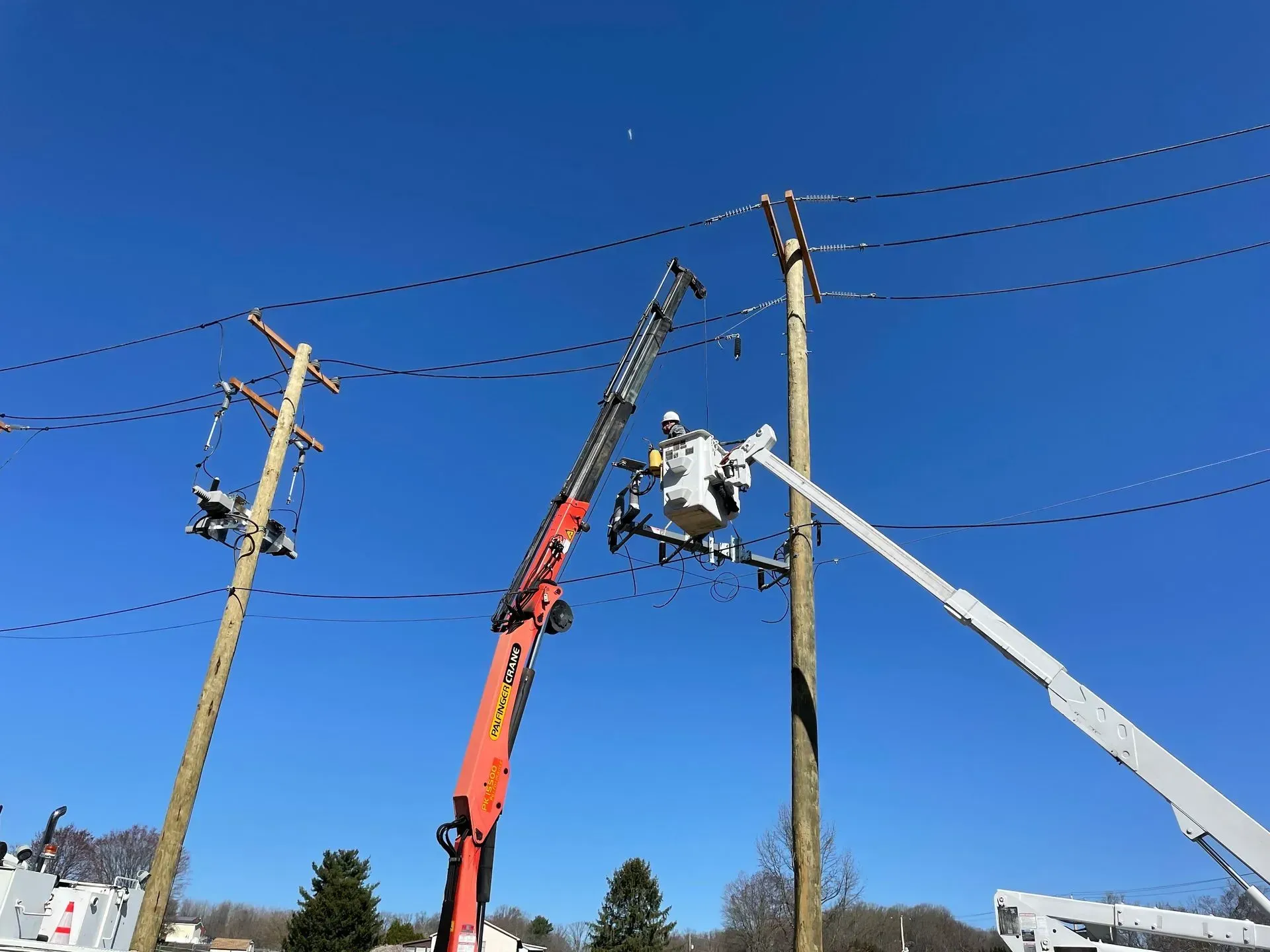 Utility workers in a bucket truck and a crane perform maintenance on power lines between two wooden utility poles.